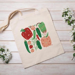 May include: A beige tote bag featuring a colorful illustration of various peppers and chili peppers. The design includes red, green, and peach-colored peppers with smiling faces and glasses. The bag has long, beige handles.