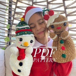 May include: A child wearing a Santa hat holds two crocheted Christmas-themed puppets. One is a snowman with a red scarf, and the other is a gingerbread man with a cherry on top. The text "PDF PATTERN" is overlaid on the image.