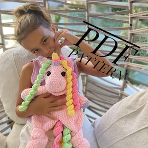 May include: A handmade pink crochet unicorn with a rainbow mane and tail. The unicorn has a smiling face and is being held. The image has the text "PDF PATTERN" across the top.