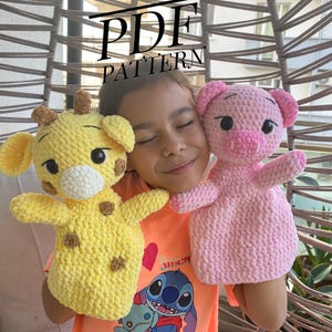 May include: A yellow crocheted giraffe and a pink crocheted pig puppet. The giraffe has brown spots and a white muzzle. The pig has a pink snout and ears. The puppets are held in front of a person wearing an orange shirt with a cartoon graphic. The text "PDF PATTERN" is visible.