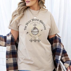 May include: Beige t-shirt featuring a graphic of the Holy Hand Grenade with a cross on top. The text around the image reads "The Holy Hand Grenade" and "To lobby at thy foe!" with the numbers "one, two, five" below. The shirt has short sleeves.