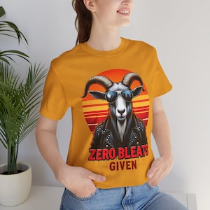Könnte beinhalten: Senfgelbes T-Shirt mit einer Ziege, die eine Sonnenbrille und eine schwarze Lederjacke trägt, vor einem roten und orangefarbenen Sonnenuntergang. Der Text "ZERO BLEATS GIVEN" ist darunter gedruckt. Ein lässiges Grafik-T-Shirt.