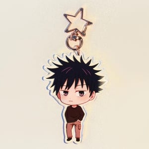 May include: A clear acrylic keychain with a cartoon character. The character has black spiky hair, a brown top, and tan trousers. The keychain has a gold-coloured star-shaped clasp.