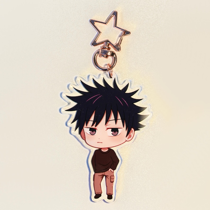 May include: A clear acrylic keychain with a cartoon character. The character has black spiky hair, a brown top, and tan trousers. The keychain has a gold-coloured star-shaped clasp.