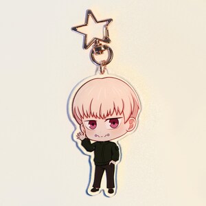 May include: A clear acrylic keychain featuring a cartoon character with pink hair and red eyes, wearing a black jacket and trousers. The keychain has a gold star-shaped clasp.