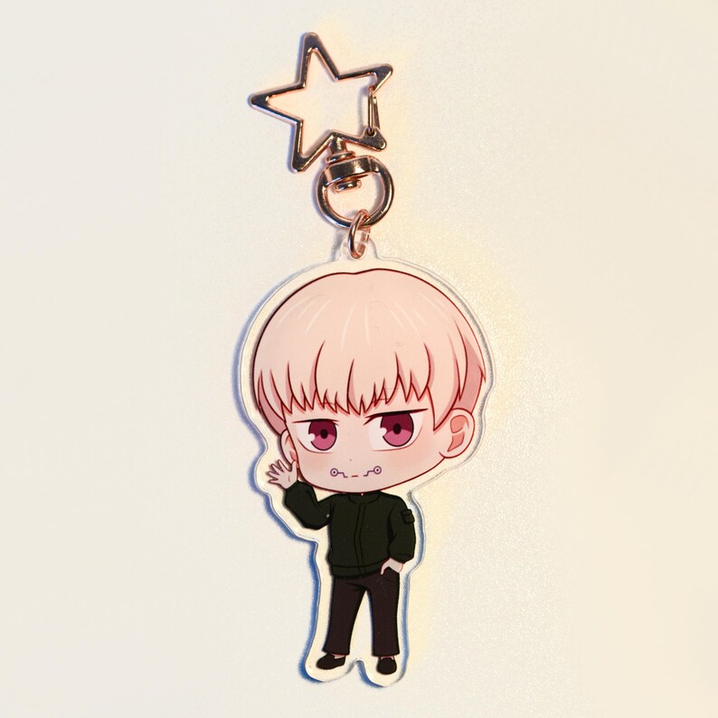 May include: A clear acrylic keychain featuring a cartoon character with pink hair and red eyes, wearing a black jacket and trousers. The keychain has a gold star-shaped clasp.