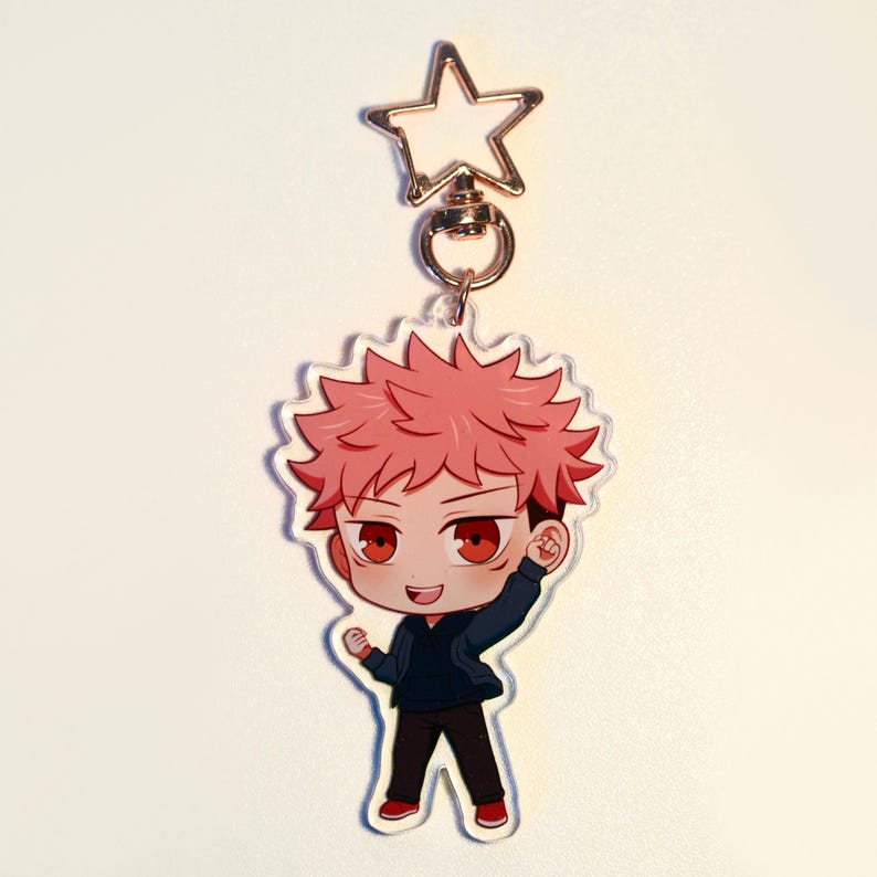 May include: A clear acrylic keychain with a cartoon character with spiky pink hair, red eyes, and a navy blue hoodie. The keychain has a gold star-shaped clasp and a gold ring. The character is smiling and has one arm raised. The character is wearing black trousers and red shoes.