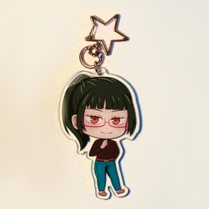 May include: A clear acrylic keychain with a cartoon character wearing glasses, a brown top, and blue trousers. The keychain has a star-shaped clasp. The character has dark hair and is wearing brown shoes.