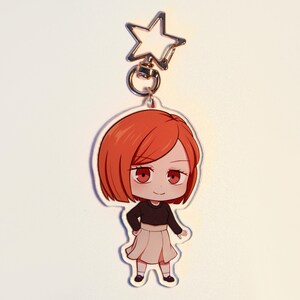 May include: A clear acrylic keychain featuring a cartoon character with red hair, red eyes, a black top, and a beige skirt. The keychain has a silver star-shaped clasp.