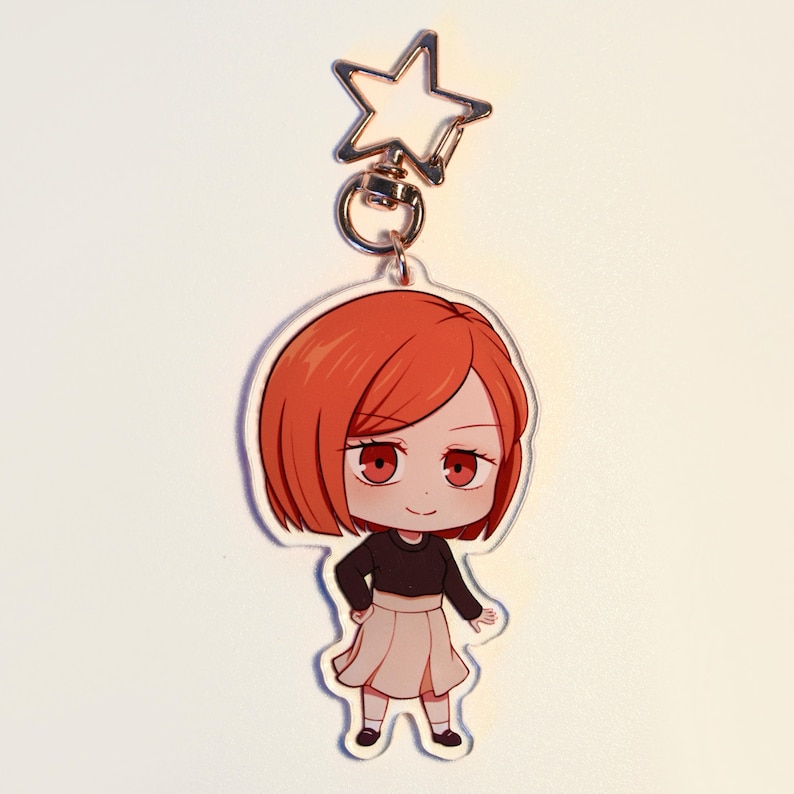 May include: A clear acrylic keychain featuring a cartoon character with red hair, red eyes, a black top, and a beige skirt. The keychain has a silver star-shaped clasp.
