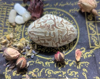 May include: An assortment of natural elements, including a patterned stone, dried flowers, and small white stones, are arranged on a dark fabric with gold lettering and symbols. The stone is the focal point, with a beige base and brown, swirling patterns.