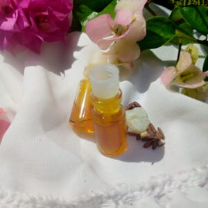 Pode incluir: Duas pequenas garrafas de vidro transparente cheias de um líquido dourado, com tampas brancas, estão sobre um pano branco. Flores rosa e magenta e folhas verdes estão no fundo, junto com algumas sementes.