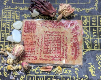 May include: A collection of dried flowers and small stones arranged around a rectangular, aged paper with red symbols and text. The background is a black cloth with gold geometric patterns and Arabic script.