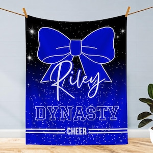 May include: A rectangular blanket featuring a black and blue gradient background with a white starfield pattern. A large blue bow is centered at the top. The text Riley is written in white script, followed by the word DYNASTY and CHEER in white.