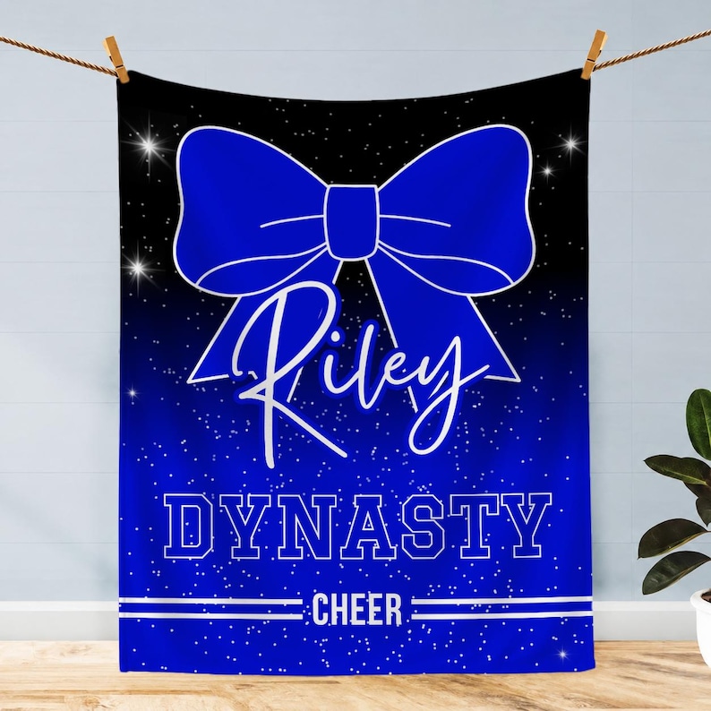May include: A rectangular blanket featuring a black and blue gradient background with a white starfield pattern. A large blue bow is centered at the top. The text Riley is written in white script, followed by the word DYNASTY and CHEER in white.