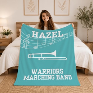 Pu&ograve; includere: Una coperta in pile color ottanio con il testo bianco HAZEL in alto, un pentagramma musicale bianco con note, un'illustrazione bianca di un trombone al centro e il testo bianco WARRIORS MARCHING BAND nella parte inferiore del tessuto.