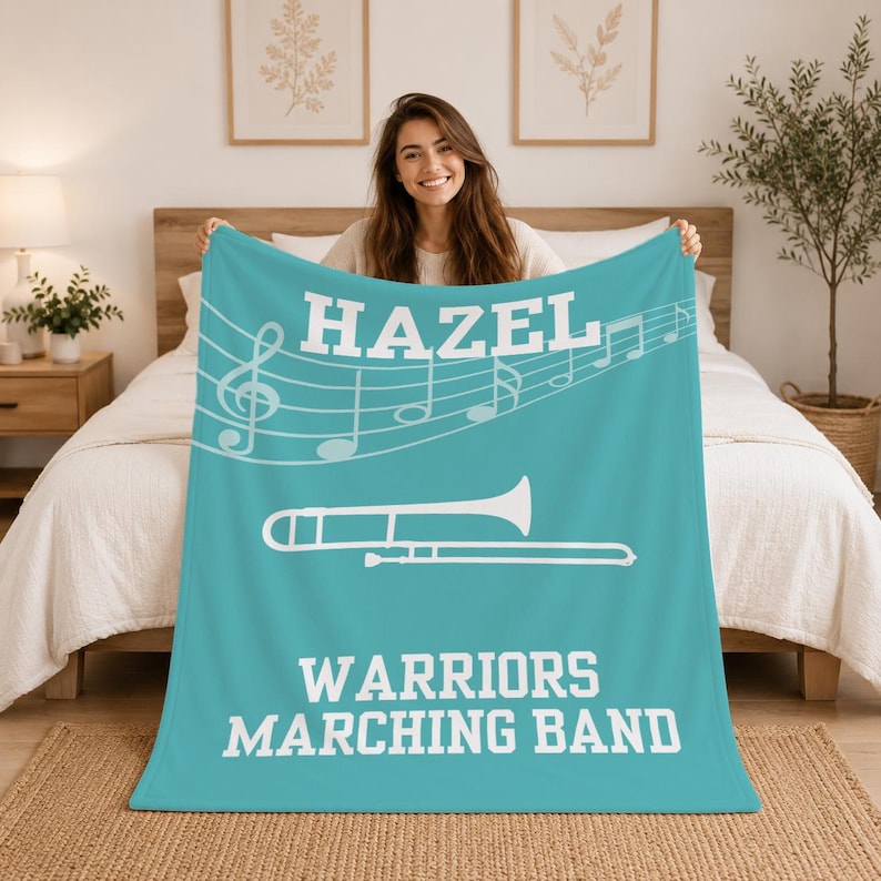 Pu&ograve; includere: Una coperta in pile color ottanio con il testo bianco HAZEL in alto, un pentagramma musicale bianco con note, un'illustrazione bianca di un trombone al centro e il testo bianco WARRIORS MARCHING BAND nella parte inferiore del tessuto.