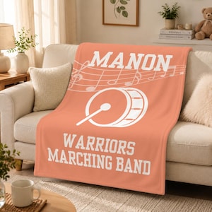Pu&ograve; includere: Una coperta in pile color corallo con il testo bianco MANON, un pentagramma musicale con note, un grafico di una grancassa con una bacchetta e il testo bianco WARRIORS MARCHING BAND stampato in un carattere a blocchi al centro della coperta.