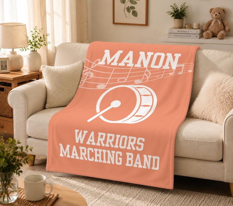 Pu&ograve; includere: Una coperta in pile color corallo con il testo bianco MANON, un pentagramma musicale con note, un grafico di una grancassa con una bacchetta e il testo bianco WARRIORS MARCHING BAND stampato in un carattere a blocchi al centro della coperta.