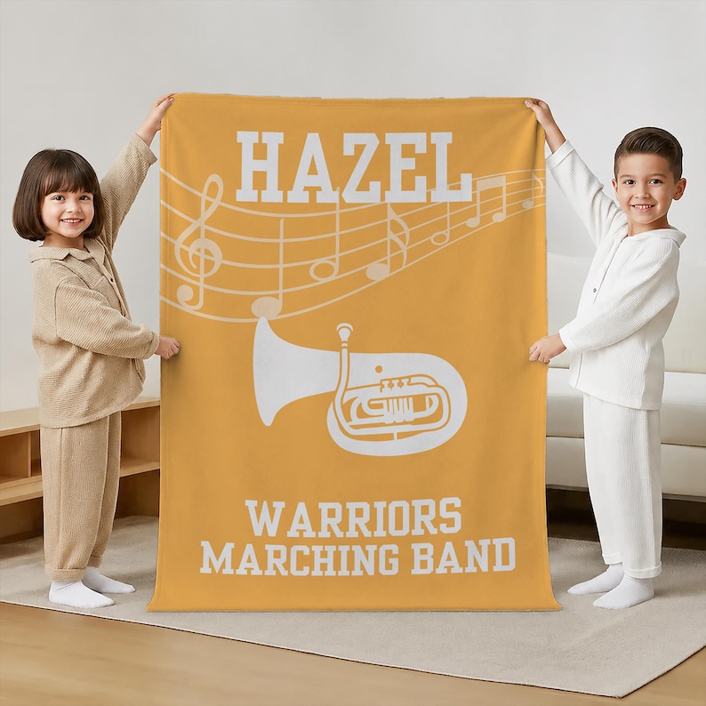 Pu&ograve; includere: Una coperta rettangolare in pile color giallo senape con il testo bianco HAZEL in alto, una grafica bianca di una tuba al centro e il testo bianco WARRIORS MARCHING BAND in basso, su uno sfondo con pentagramma e note musicali bianche.