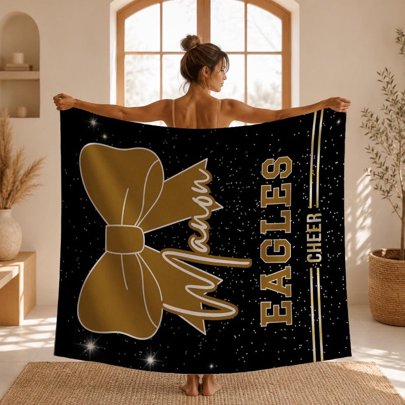 May include: A black blanket featuring a large gold bow graphic, the name Mason in script, and the text EAGLES CHEER in gold block letters. The design includes a gold vertical line and a scattered white star pattern across the dark background.