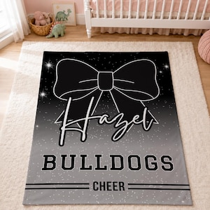 May include: A rectangular blanket featuring a black and grey gradient background with a white speckled star pattern. A large black bow graphic sits at the top. The name Hazel is written in white script, followed by the text BULLDOGS and CHEER.