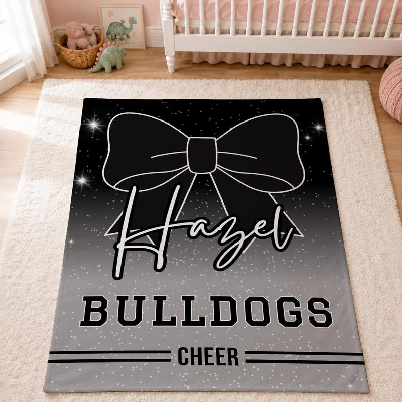 May include: A rectangular blanket featuring a black and grey gradient background with a white speckled star pattern. A large black bow graphic sits at the top. The name Hazel is written in white script, followed by the text BULLDOGS and CHEER.