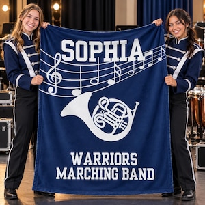Pu&ograve; includere: Una coperta in pile blu scuro con il testo bianco SOPHIA in alto, un pentagramma musicale bianco con note, una grafica bianca di un corno al centro e il testo bianco WARRIORS MARCHING BAND stampato nella parte inferiore del tessuto.