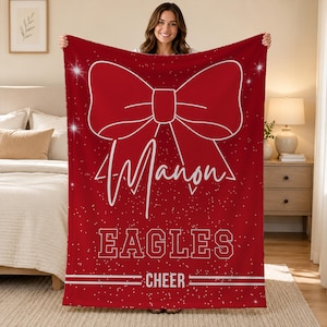 May include: A rectangular red fleece blanket featuring a large white outline of a bow at the top. Below the bow, the name Manon is printed in white script, followed by the word EAGLES in block letters and the word CHEER with horizontal lines.