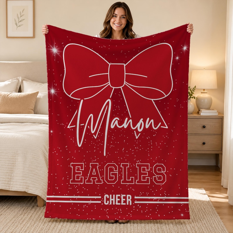 May include: A rectangular red fleece blanket featuring a large white outline of a bow at the top. Below the bow, the name Manon is printed in white script, followed by the word EAGLES in block letters and the word CHEER with horizontal lines.
