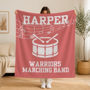 Pu&ograve; includere: Una coperta in pile color corallo con la scritta bianca HARPER in alto, una grafica bianca di un rullante con due bacchette e la scritta bianca WARRIORS MARCHING BAND in basso, arricchita da un pentagramma e note musicali bianche.