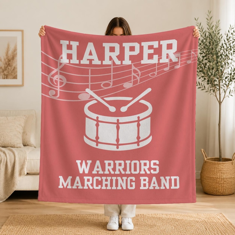 Pu&ograve; includere: Una coperta in pile color corallo con la scritta bianca HARPER in alto, una grafica bianca di un rullante con due bacchette e la scritta bianca WARRIORS MARCHING BAND in basso, arricchita da un pentagramma e note musicali bianche.