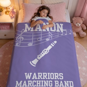 Pu&ograve; includere: Una coperta rettangolare in pile viola con una grafica bianca di un clarinetto, un pentagramma e note musicali. Il testo recita MANON in alto e WARRIORS MARCHING BAND in basso in grassetto con lettere maiuscole bianche ben visibili.