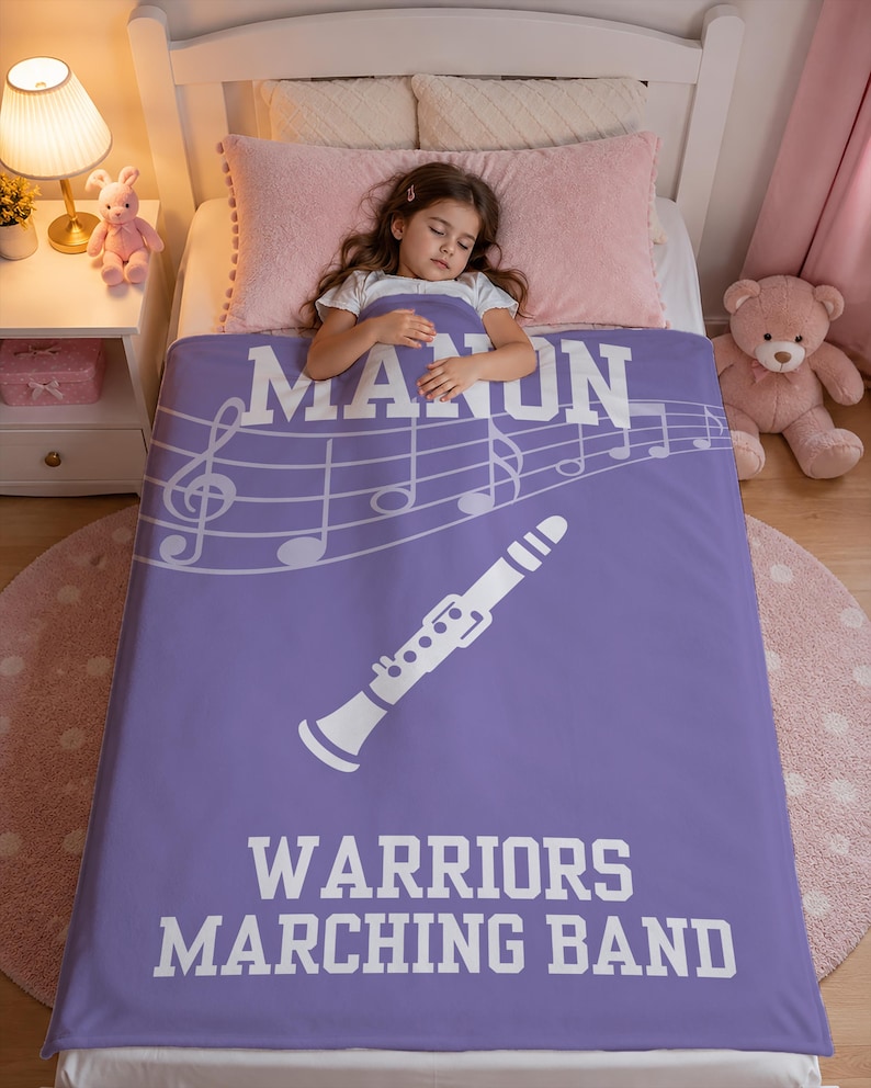 Pu&ograve; includere: Una coperta rettangolare in pile viola con una grafica bianca di un clarinetto, un pentagramma e note musicali. Il testo recita MANON in alto e WARRIORS MARCHING BAND in basso in grassetto con lettere maiuscole bianche ben visibili.