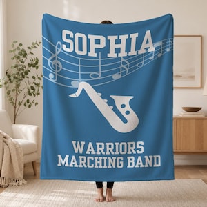 Pu&ograve; includere: Una coperta in pile blu con il testo bianco SOPHIA in alto, un grafico bianco di un sassofono al centro e il testo bianco WARRIORS MARCHING BAND in basso, con un pentagramma e note musicali bianche dietro il nome.