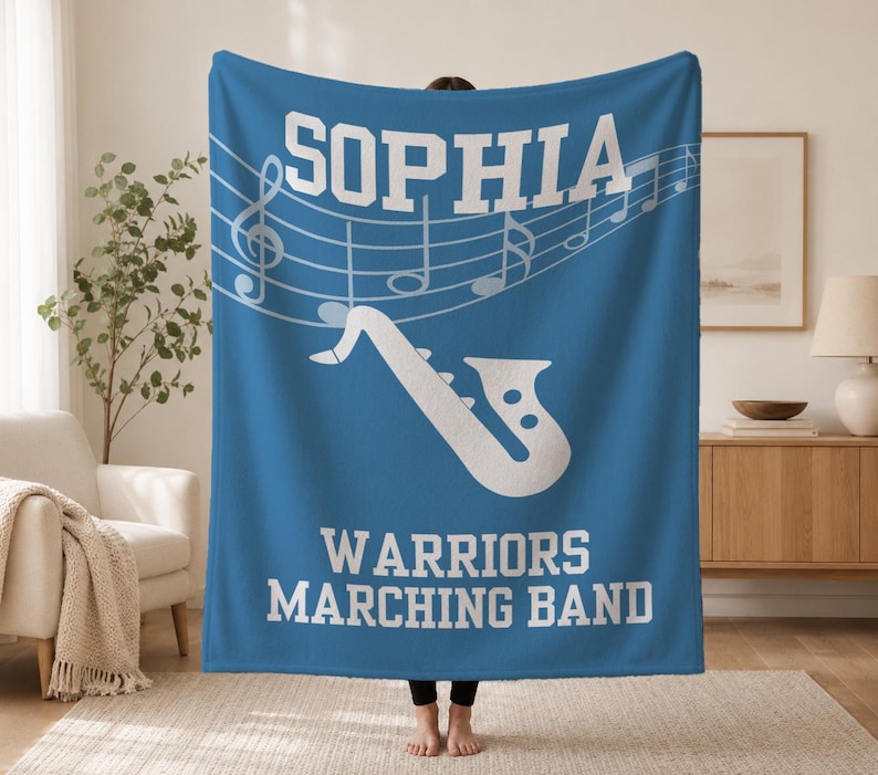 Pu&ograve; includere: Una coperta in pile blu con il testo bianco SOPHIA in alto, un grafico bianco di un sassofono al centro e il testo bianco WARRIORS MARCHING BAND in basso, con un pentagramma e note musicali bianche dietro il nome.