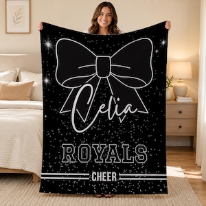 May include: A black rectangular fleece blanket featuring a large white outline of a bow at the top. The name Celia is printed in white script below the bow. The text ROYALS is in white block letters, followed by the word CHEER and white lines.