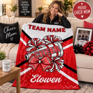 May include: A white and red fleece blanket featuring the text TEAM NAME and Elowen, with a graphic of a megaphone, a large bow, and pom-poms. The design includes black diagonal stripes and a glittery red texture on the cheer-themed elements.