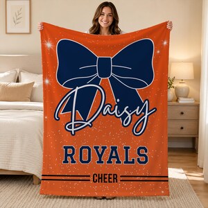 May include: An orange fleece blanket featuring a large dark blue bow graphic, white star accents, and the text Daisy, ROYALS, and CHEER in dark blue lettering. The design includes horizontal lines framing the word CHEER at the bottom of the blanket.