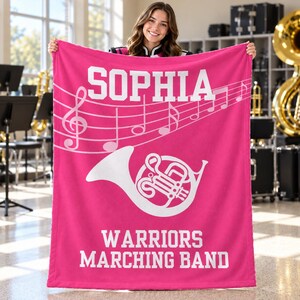 Pu&ograve; includere: Una coperta in pile rosa acceso con il testo bianco SOPHIA in alto, un pentagramma musicale bianco con note, una grafica bianca di un corno al centro e il testo bianco WARRIORS MARCHING BAND nella parte inferiore del tessuto rettangolare.