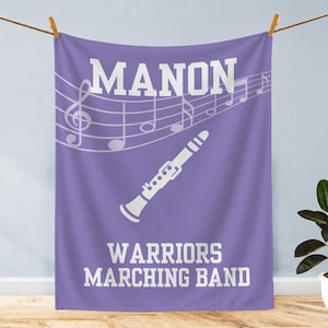 Pu&ograve; includere: Una coperta rettangolare viola con una grafica bianca di un clarinetto, linee di pentagramma con note musicali e il testo bianco MANON WARRIORS MARCHING BAND stampato in un carattere in grassetto nella parte superiore e inferiore.