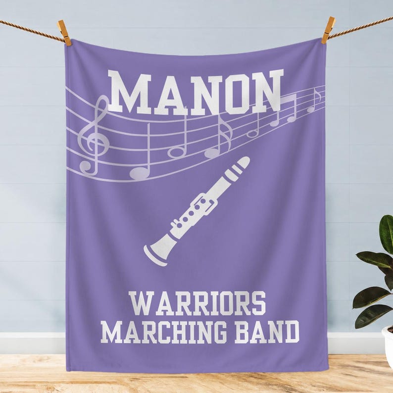 Pu&ograve; includere: Una coperta rettangolare viola con una grafica bianca di un clarinetto, linee di pentagramma con note musicali e il testo bianco MANON WARRIORS MARCHING BAND stampato in un carattere in grassetto nella parte superiore e inferiore.