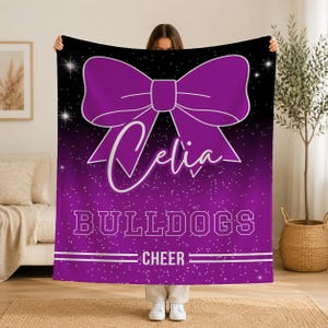 May include: A rectangular fleece blanket featuring a black and purple gradient background with a white glitter effect. A large purple bow is centered at the top. The name Celia is written in white script, followed by the text BULLDOGS and CHEER.
