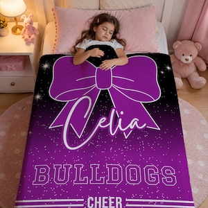 May include: A rectangular blanket featuring a purple and black gradient background with a white glitter effect. The design includes a large purple bow, the name Celia in white script, the word BULLDOGS in block letters, and the word CHEER.