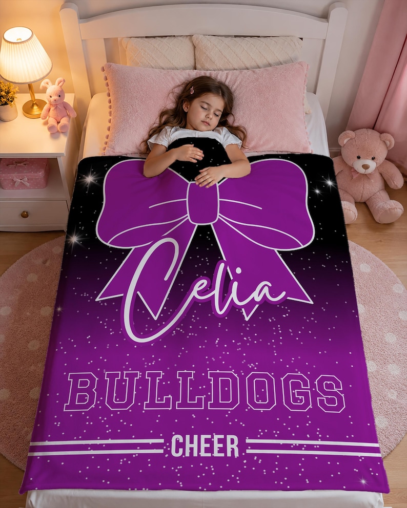 May include: A rectangular blanket featuring a purple and black gradient background with a white glitter effect. The design includes a large purple bow, the name Celia in white script, the word BULLDOGS in block letters, and the word CHEER.