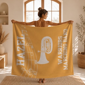 Pu&ograve; includere: Una coperta color giallo senape con una grafica bianca di una tuba, note musicali su un pentagramma e il testo "HAZEL" sul lato sinistro e "WARRIORS MARCHING BAND" sul lato destro in un carattere bianco, grassetto e senza grazie.