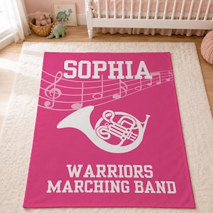Pu&ograve; includere: Una coperta rettangolare in pile rosa con il testo bianco SOPHIA in alto, un pentagramma musicale bianco con note al centro, una grafica bianca di un corno e il testo bianco WARRIORS MARCHING BAND nella parte inferiore della coperta.