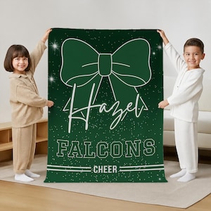 May include: A dark green rectangular blanket featuring a large white graphic of a hair bow at the top. The text "Hazel" is written in a white script font below the bow, followed by the block text "FALCONS" and "CHEER" with white sparkles.