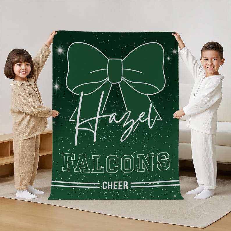 May include: A dark green rectangular blanket featuring a large white graphic of a hair bow at the top. The text "Hazel" is written in a white script font below the bow, followed by the block text "FALCONS" and "CHEER" with white sparkles.