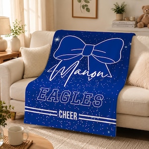 May include: A royal blue fleece blanket featuring a large white outline of a hair bow at the top, the name Manon in white script, the word EAGLES in white block letters, and the word CHEER in white text between two horizontal white lines.